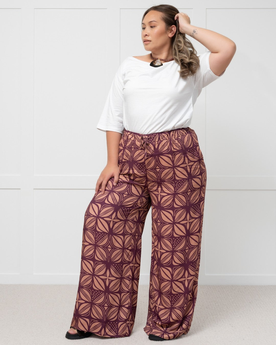 Woman wearing a white top and patterned purple island pants against a white background