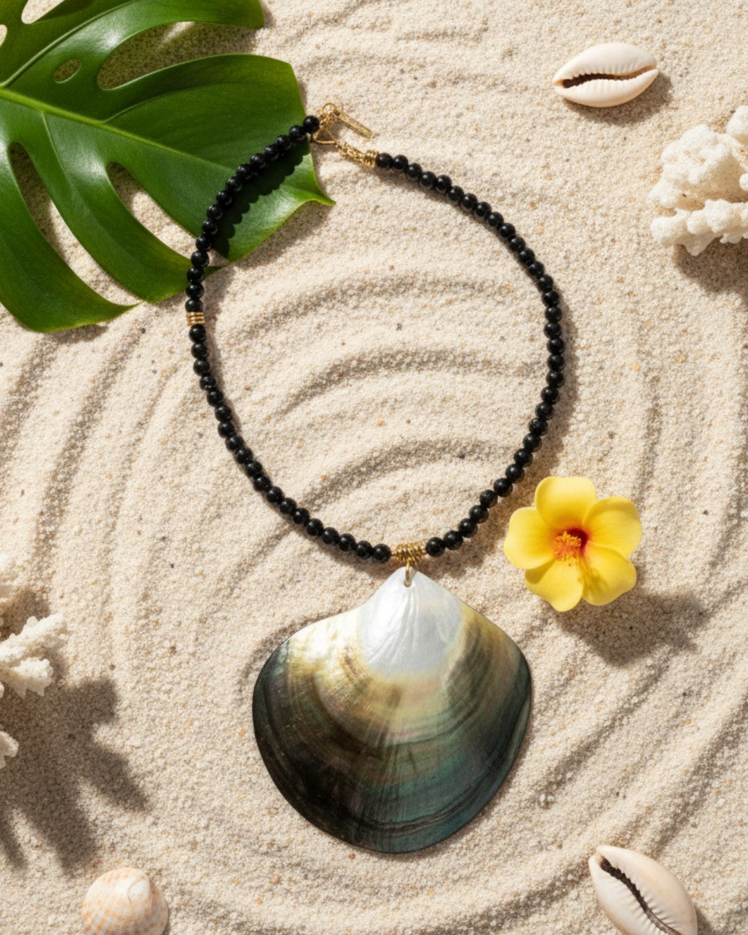 Necklace with black beads and a yellow flower on a shell, placed on sand with leaves and shells around.