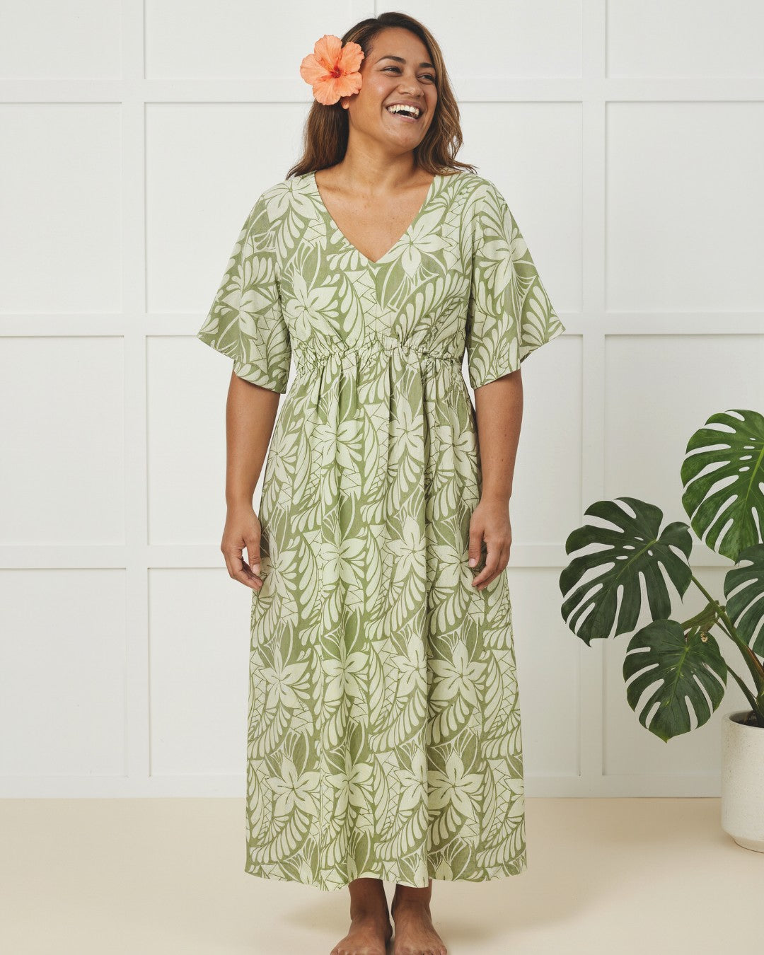 Woman wearing a green leaf-patterned dress with a white wall and plant in the background