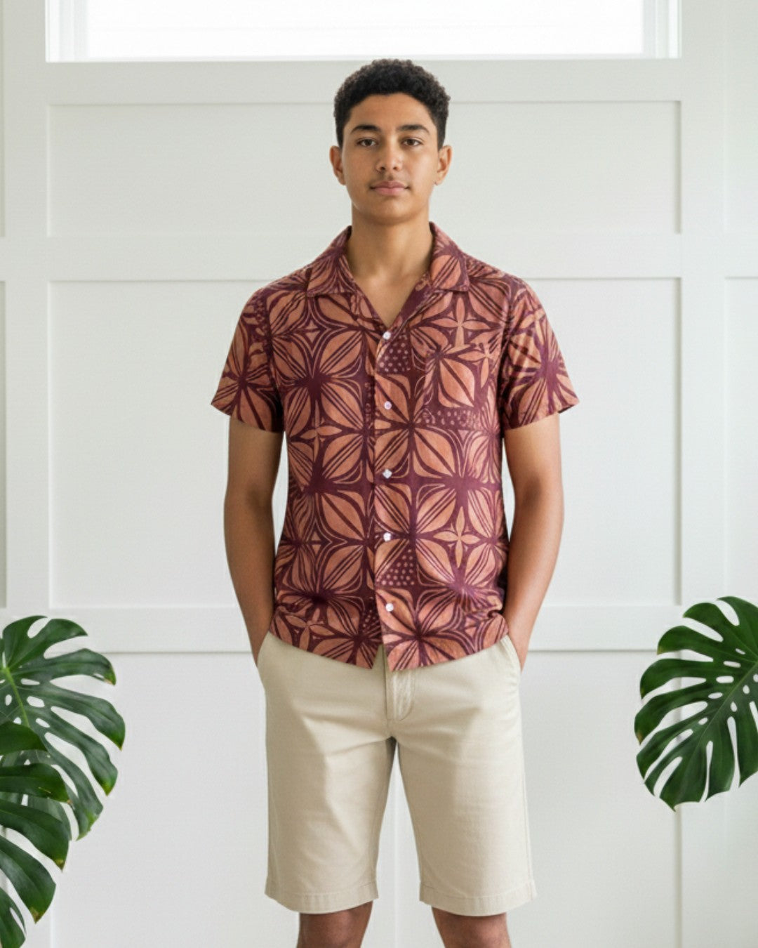 Person wearing a patterned shirt and beige shorts standing against a white paneled wall with green leaves.