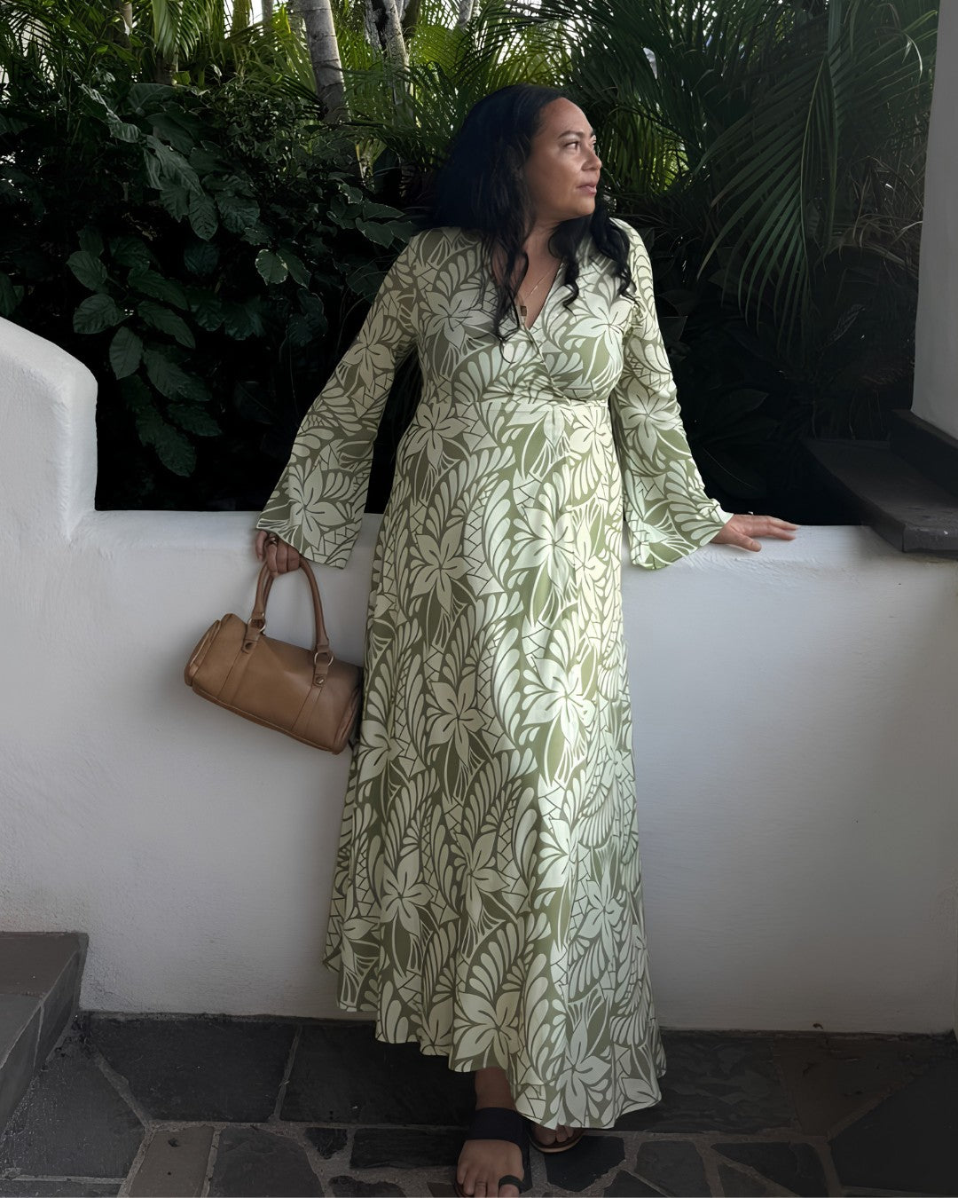 Woman in a green leaf-patterned dress holding a brown handbag against a white wall with greenery.