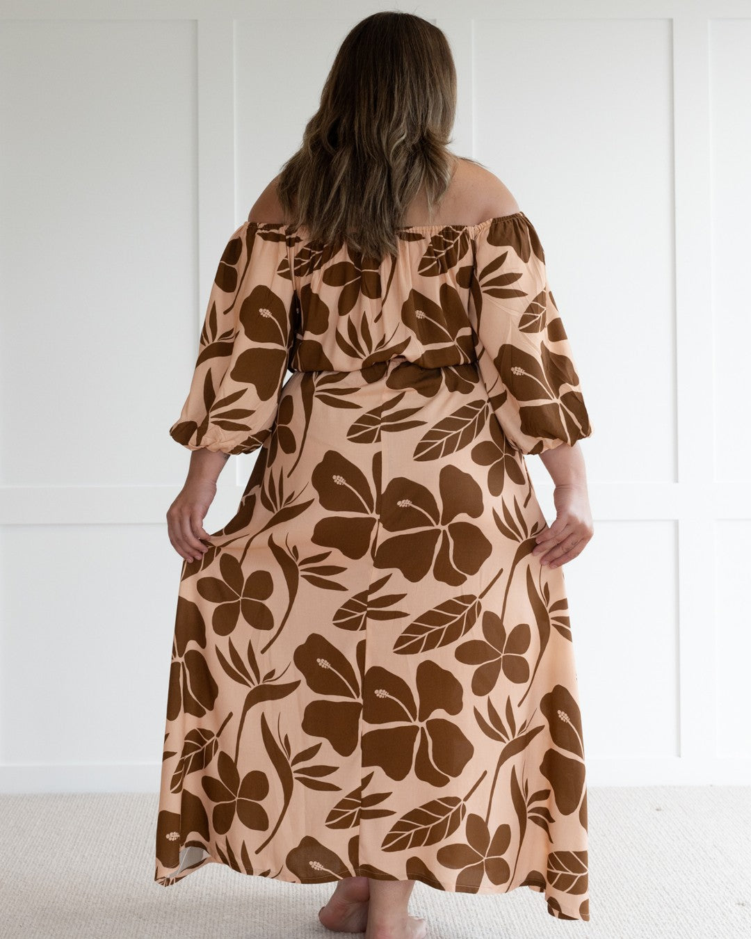 Woman wearing a floral off-shoulder island dress against a white wall.
